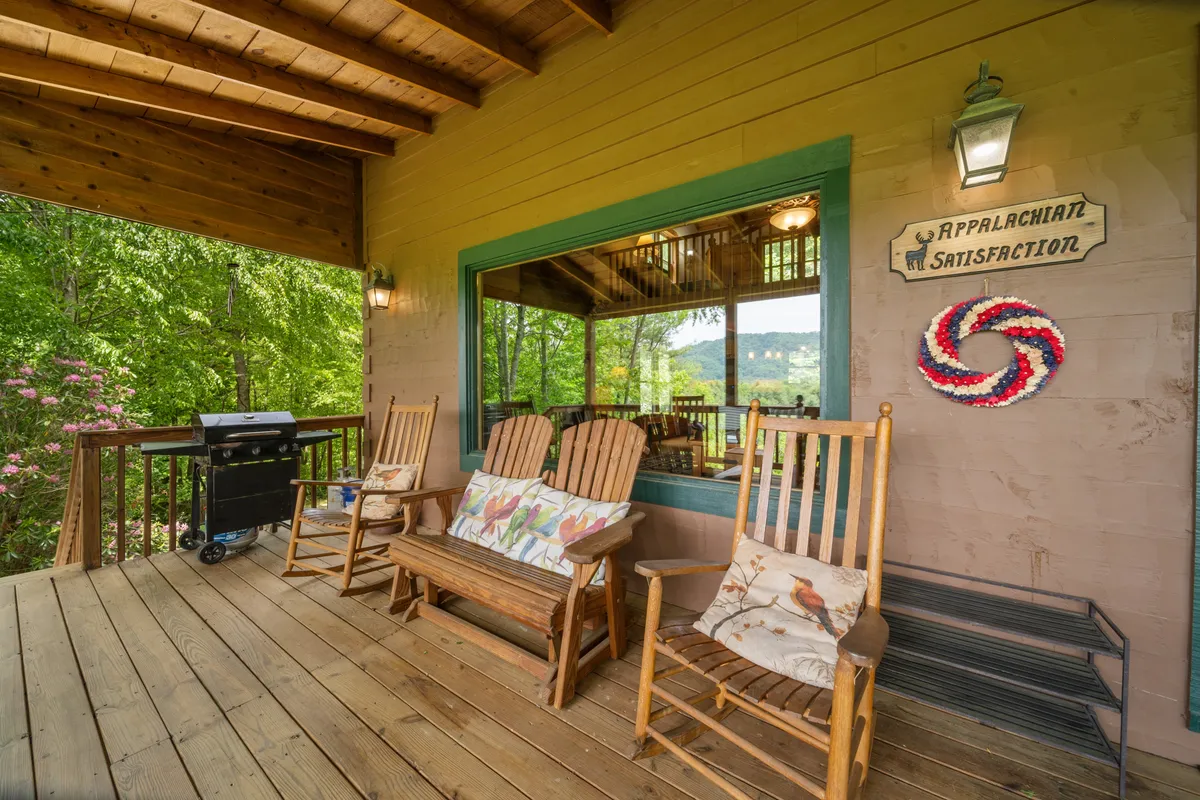 A cozy porch scene featuring rocking chairs, a barbecue grill, a large window, and a decorative sign. The landscaped view outside adds to the setting's inviting ambiance, ideal for outdoor meals and gatherings.