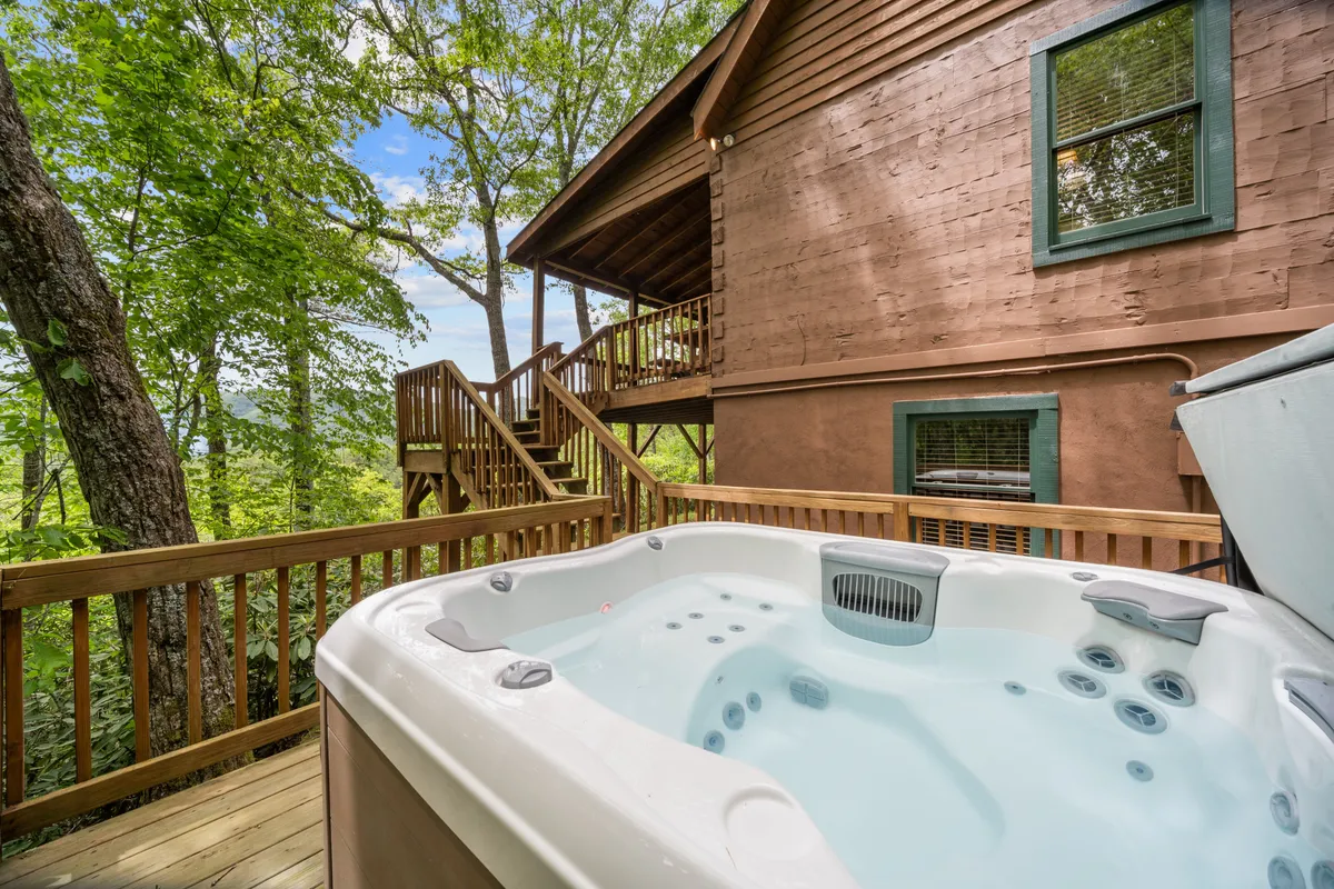 The image showcases the exterior of a classic log cabin nestled among tall trees, featuring a spacious wooden deck with a bubbling hot tub. The scene evokes relaxation, with the deck offering panoramic sunset views, perfect for morning coffee or evening stargazing, and surrounded by tranquil nature.