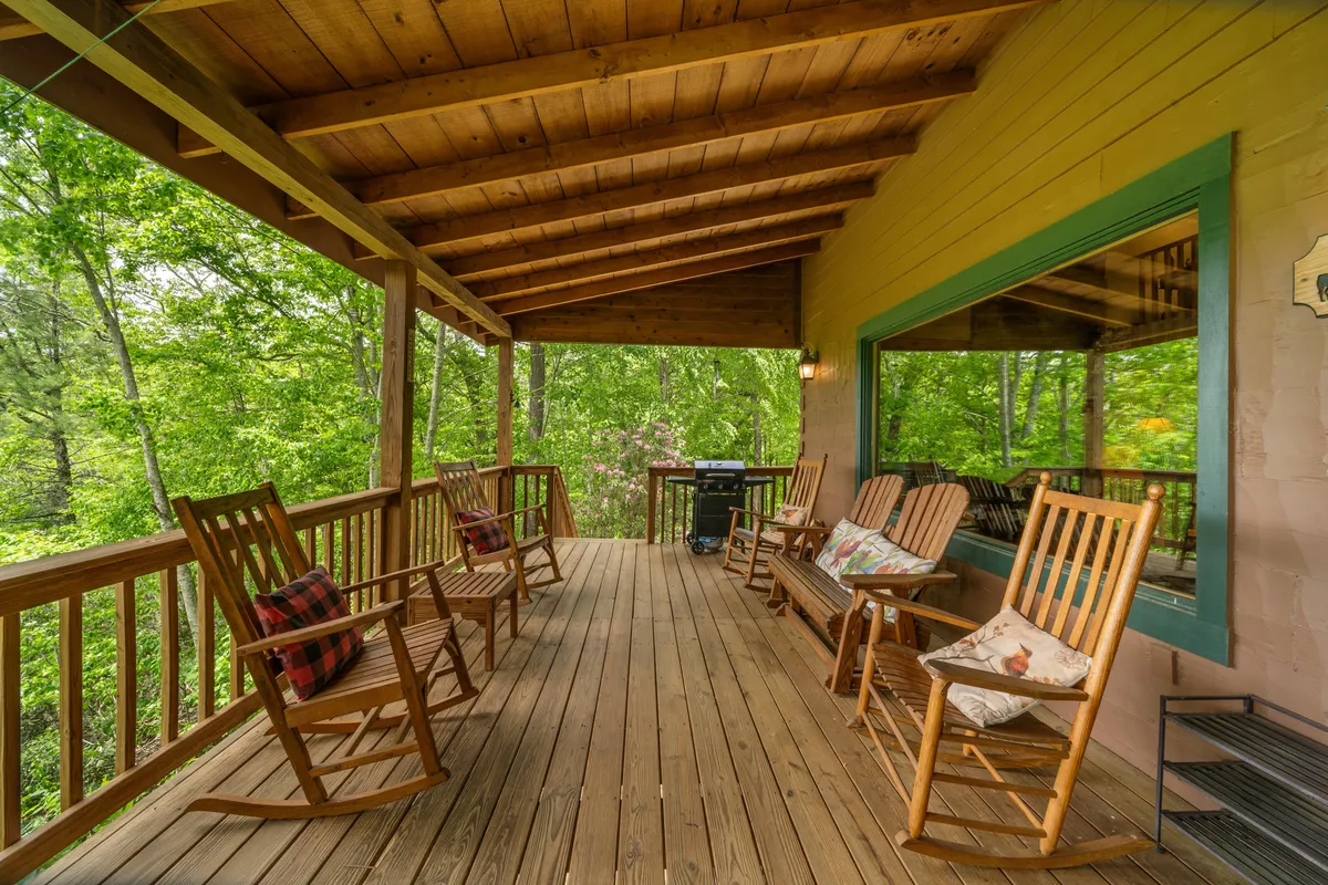 A welcoming exterior shot featuring a deck with several rocking chairs, a wooden railing, surrounding greenery, a barbecue grill, and a large window that hints at the inviting interior. The deck offers ample space for group relaxation, outdoor meals, and socializing while enjoying nature.