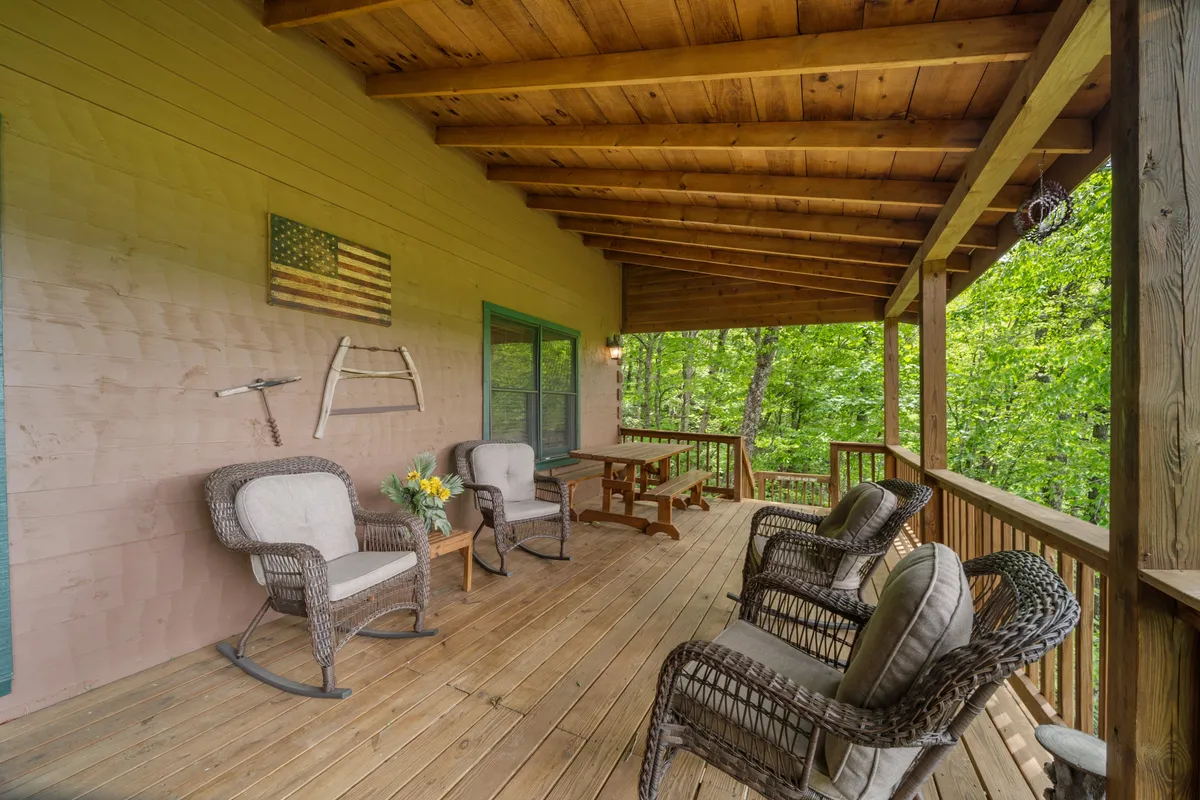 An inviting exterior porch featuring wooden rocking chairs, a wooden table, and an American flag motif, all set against a backdrop of natural scenery. The setup encourages relaxation, socializing, and taking in the quiet surroundings.