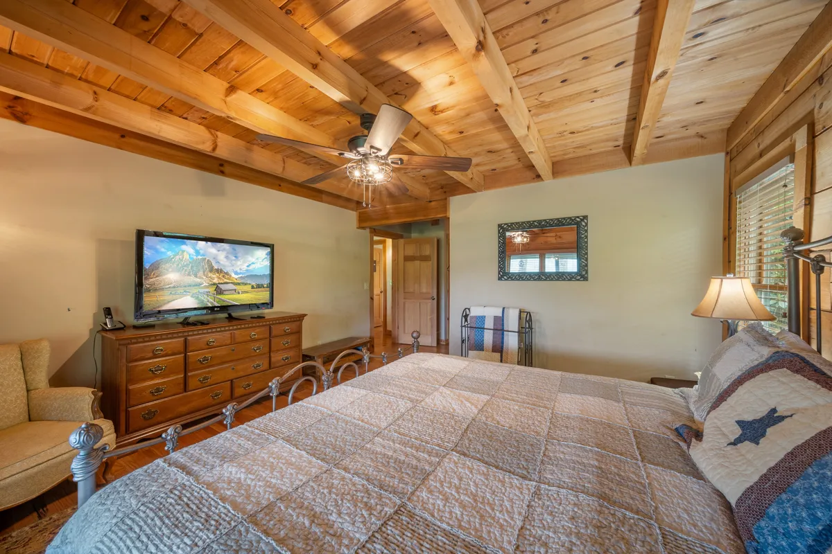 A warmly lit bedroom featuring a king-sized bed, a wooden dresser, a television, and a cozy sitting area with an armchair. The room exudes an inviting and restful atmosphere, ideal for relaxation and privacy.