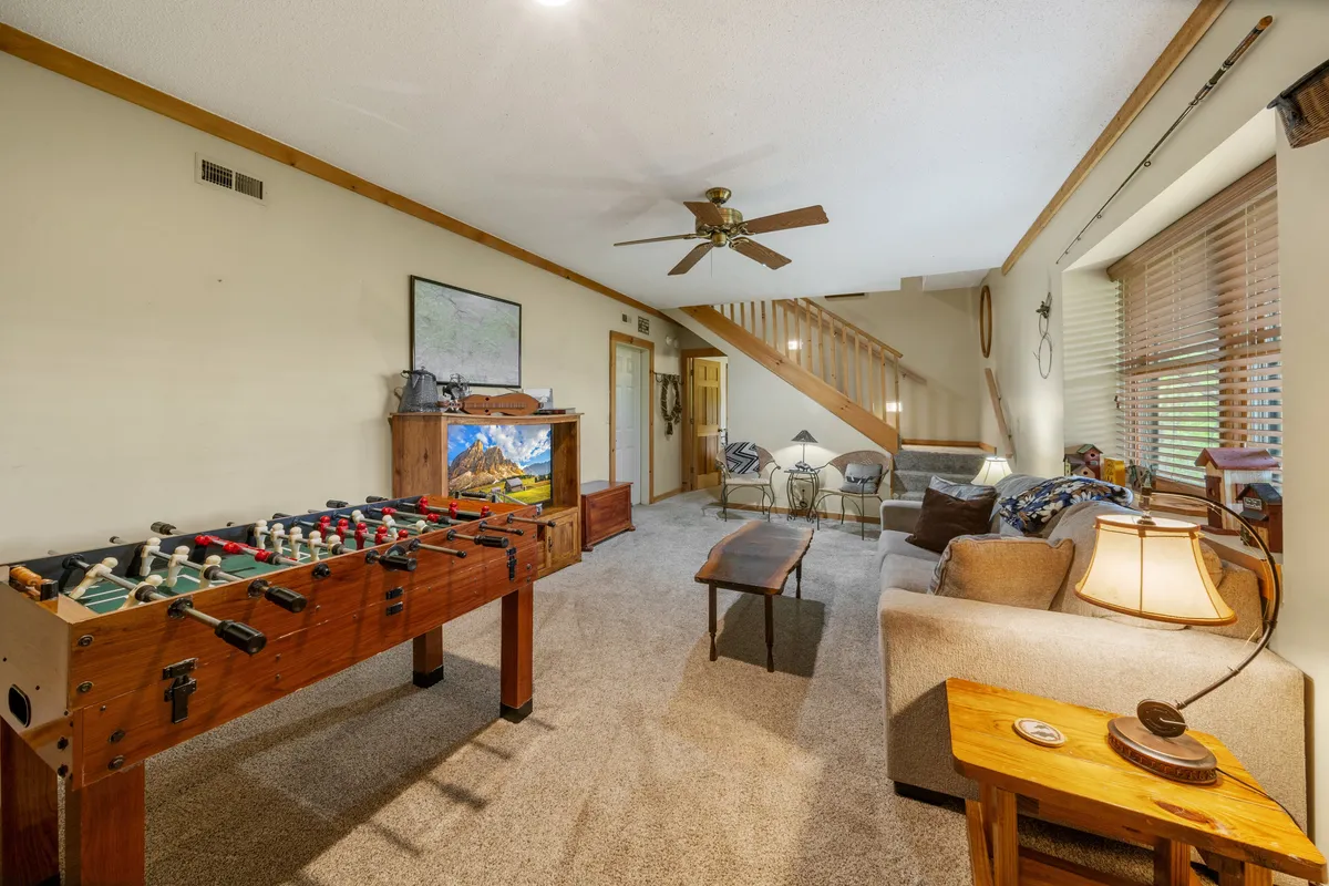 This image features a cozy lower-level family room with a foosball table, a comfortable sofa, a wooden coffee table, and a TV stand with a picture of the mountains. The space feels inviting and playful, offering both relaxation and opportunities for games and shared fun.