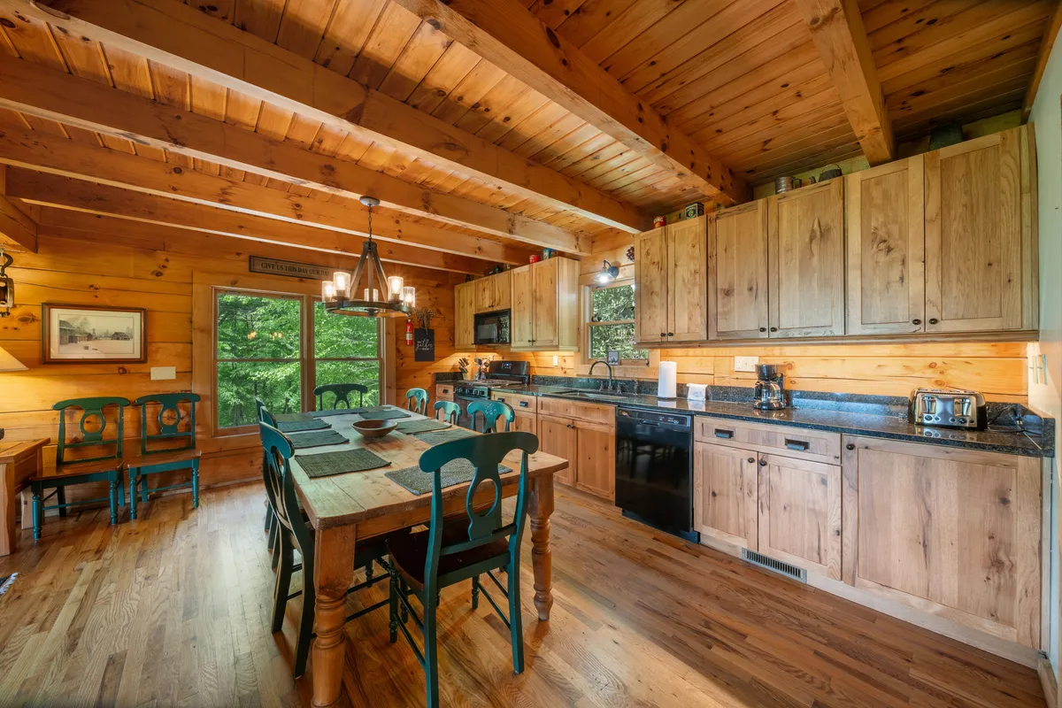 The focus is on a large wooden dining table adjacent to the kitchen, with windows framing greenery outside and a chandelier overhead. The table is ready for family meals, games, or celebrations, while the kitchen cabinetry and countertops hint at a fully equipped, functional space.