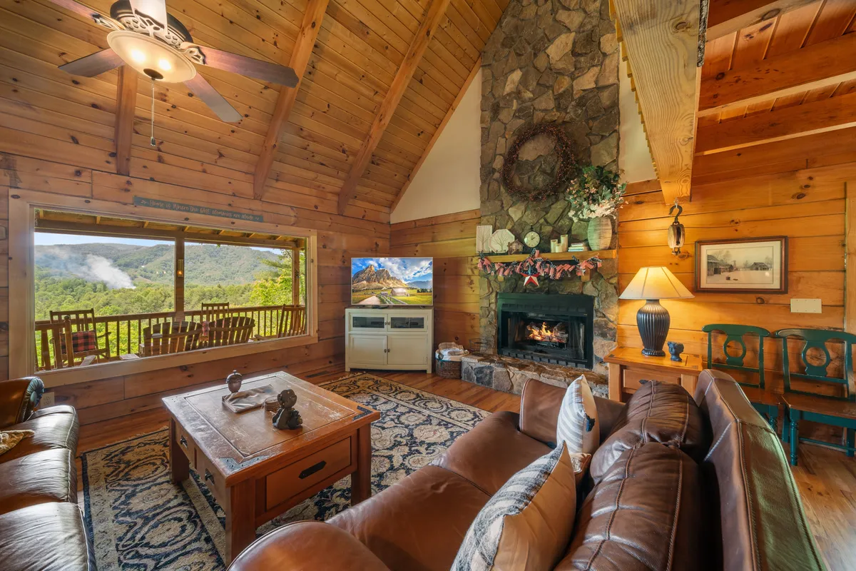 This image features a large stone fireplace as the stunning centerpiece of a spacious living area. The room is filled with natural light from an expansive window that frames a beautiful mountain view. A comfortable leather sofa and a decorative wooden coffee table invite relaxation, while a television with a nature scene enhances the cozy atmosphere.